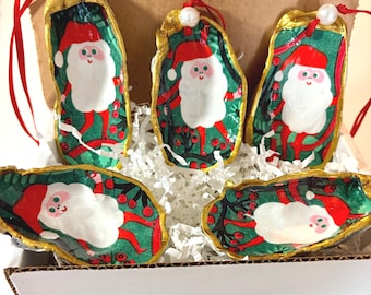 May include: Five oyster shell ornaments featuring hand-painted Santa Claus figures with white beards and red hats against a green background with red berries. Each shell has a gold-painted edge, a white bead, and a red ribbon for hanging.
