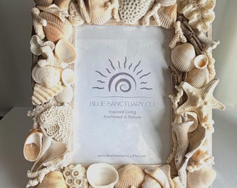 May include: A rectangular picture frame decorated with various white and tan seashells, coral pieces, and driftwood. The center insert features the text BLUE SANCTUARY CO. Inspired Living Anchored in Nature and the website www.BlueSanctuaryCo.Etsy.com.
