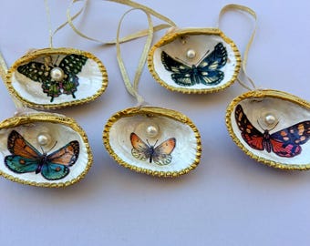 May include: Five decorative seashell ornaments, each featuring a unique butterfly illustration inside, a central white pearl accent, a gold-painted scalloped edge, and a thin gold ribbon loop for hanging against a plain light background.