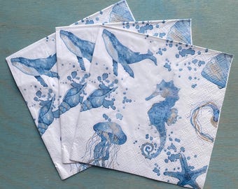 Puede incluir: Un juego de servilletas de papel cuadradas con un estampado estilo acuarela de vida marina azul, que incluye ballenas, un caballito de mar, una medusa, una estrella de mar y conchas marinas, sobre un fondo blanco con salpicaduras de pintura.