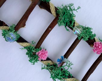 May include: A decorative wooden ladder adorned with vibrant green moss, small pink rose-shaped embellishments, shimmering gold ribbon wrapped around the rungs, clear iridescent spherical beads, and a tiny blue butterfly accent piece.