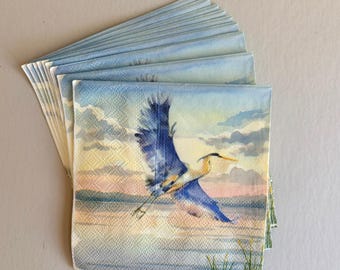 Puede incluir: Una pila de servilletas de papel con una pintura a la acuarela de una garza azul en vuelo sobre una masa de agua. El fondo presenta una puesta de sol con tonos rosas, amarillos y azules. Las servilletas son de color azul claro.