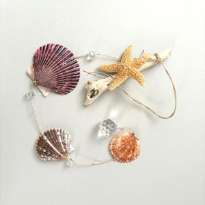 May include: A decorative hanging ornament featuring a piece of light-colored driftwood, a textured orange starfish, three assorted seashells in shades of purple and brown, and clear faceted crystal beads strung together on thin natural twine.