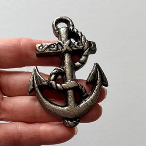May include: A metallic silver-toned anchor pendant featuring a textured, weathered finish. The design includes a rope wrapped around the central shank and intricate scrollwork detailing on the horizontal crossbar, with a loop at the top for hanging.