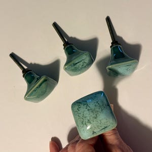 May include: Four square cabinet knobs with a glossy, mottled teal and turquoise glaze. Each knob features a smooth, rounded-edge square face and a threaded metal bolt extending from the back for installation, showcasing a unique speckled finish.