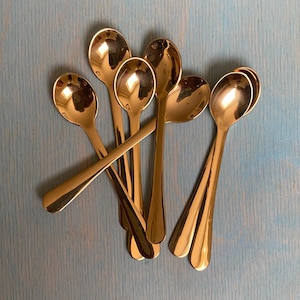 May include: A collection of shiny, gold-colored spoons arranged on a light blue surface. The spoons have a classic design with rounded bowls and long, slender handles. The spoons are reflective, catching the light and creating highlights.