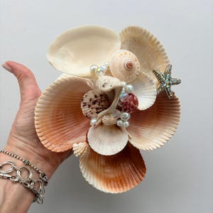 May include: A decorative arrangement of various seashells including fan-shaped scallops, spiral shells, and a small starfish with iridescent crystals. The cluster is accented with white spherical beads and clear faceted glass beads.
