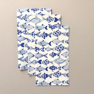 May include: Three white paper napkins with a repeating pattern of blue fish. The fish designs vary, including stripes, dots, and solid blue. The napkins are stacked slightly, showing the pattern on each.