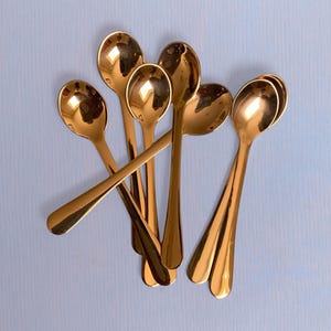 May include: A collection of six polished copper-toned metal spoons arranged in a cluster. Each spoon features a smooth, reflective surface with a rounded bowl and a slender handle, showcasing a uniform metallic finish throughout the set.