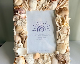 May include: A rectangular picture frame decorated with a variety of natural seashells and coral pieces. The center insert features the purple logo of Blue Sanctuary Co. with the text Inspired Living Anchored in Nature and the website address.