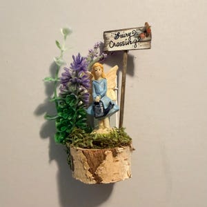 May include: A miniature figurine of a blonde fairy in a blue dress holding a black watering can, standing on a birch wood base with green moss, purple flowers, and a wooden signpost that reads Fairy Crossing with a small orange butterfly.