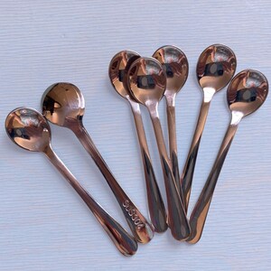 May include: A set of six polished rose gold-toned metal spoons with rounded bowls and slender handles. Each spoon features a smooth, reflective finish, and one handle is clearly engraved with the text SUS304, indicating the stainless steel grade.