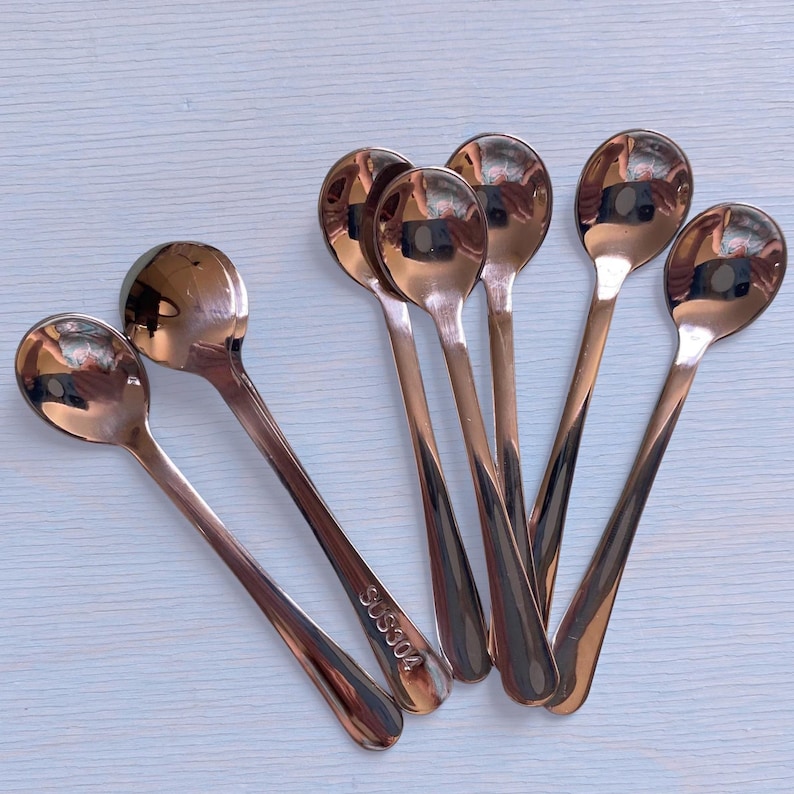 May include: A set of six polished rose gold-toned metal spoons with rounded bowls and slender handles. Each spoon features a smooth, reflective finish, and one handle is clearly engraved with the text SUS304, indicating the stainless steel grade.