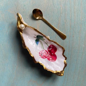 May include: A decorative oyster shell dish with a white interior featuring a painted red cherry design, gold-painted edges, and a single white pearl accent. A small, polished gold-tone metal spoon rests beside the shell on a light blue surface.