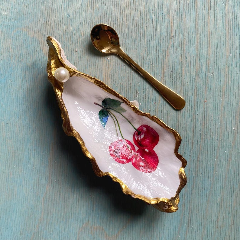 May include: A decorative oyster shell dish with a white interior featuring a painted red cherry design, gold-painted edges, and a single white pearl accent. A small, polished gold-tone metal spoon rests beside the shell on a light blue surface.