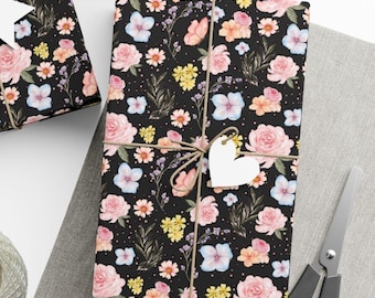 May include: A rectangular gift wrapped in black paper featuring a floral pattern of pink roses, light blue hydrangeas, yellow wildflowers, and butterflies. The package is tied with brown twine and includes a blank white heart-shaped tag.