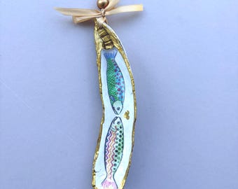 May include: A decorative oyster shell ornament featuring a hand-painted design of two fish with colorful patterns. The shell edges are painted gold, and it hangs from a light beige satin ribbon tied in a bow with a small gold bead accent.