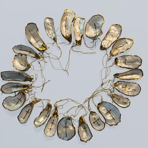 Puede incluir: Un conjunto de veinti&uacute;n adornos individuales de concha de ostra dispuestos en un patr&oacute;n circular. Cada concha presenta una cruz pintada en oro en el interior, bordes dorados y una peque&ntilde;a cuenta de perla blanca unida a un fino cord&oacute;n dorado.