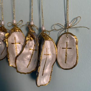 Puede incluir: Una colecci&oacute;n de adornos colgantes de conchas de ostra, pintadas de blanco con una cruz dorada en el centro y bordes dorados. Cada concha cuelga de un cord&oacute;n met&aacute;lico dorado con una peque&ntilde;a perla blanca y un lazo decorativo dorado.