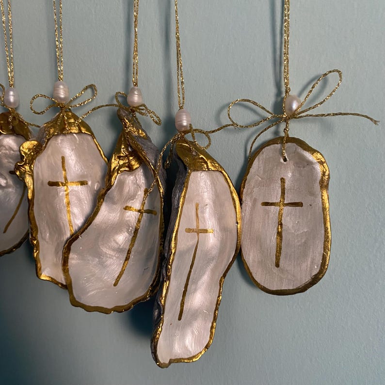 Puede incluir: Una colecci&oacute;n de adornos colgantes de conchas de ostra, pintadas de blanco con una cruz dorada en el centro y bordes dorados. Cada concha cuelga de un cord&oacute;n met&aacute;lico dorado con una peque&ntilde;a perla blanca y un lazo decorativo dorado.