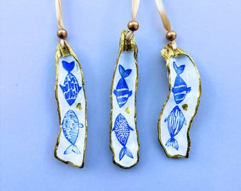 May include: Three hanging ornaments made from oyster shells with gold-painted edges. Each shell features two hand-painted blue fish illustrations and a small gold heart. A gold bead and a light beige ribbon are attached to the top of each.