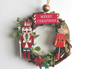 May include: A small circular wreath made of brown twigs, decorated with green leaves, a red poinsettia, a nutcracker figure, and a mouse soldier figure. A red sign with white text reads MERRY CHRISTMAS, topped with a gold bead and ribbon.