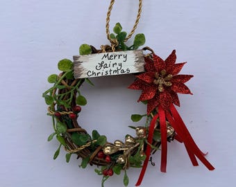 Puede incluir: Un pequeño adorno de corona circular hecho de ramas marrones, follaje verde y bayas rojas. Incluye un letrero de madera rectangular con el texto Merry Fairy Christmas, una flor de pascua roja brillante, cintas rojas y un cordón dorado.