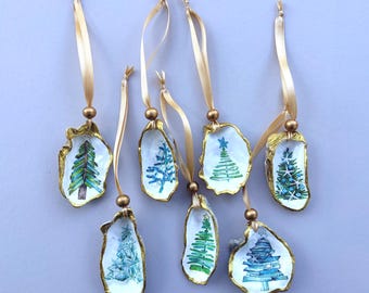 May include: Seven oyster shell ornaments featuring hand-painted green and blue Christmas tree designs. Each shell has a gold-painted edge and is suspended by a gold ribbon with a small gold bead at the top for hanging on a tree.