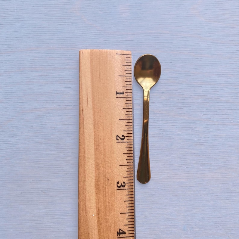 May include: A small gold-toned metal spoon positioned next to a wooden ruler. The spoon measures approximately 3.5 inches in total length, featuring a smooth, reflective surface, a rounded bowl, and a slender handle that tapers toward the end.