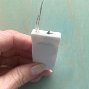 May include: A rectangular white plastic battery pack for LED string lights, featuring a small black sliding power switch on the top edge and two thin, clear electrical wires extending upward from the center of the housing unit.