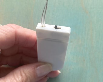 May include: A rectangular white plastic battery pack for LED string lights, featuring a small black sliding power switch on the top edge and two thin, clear electrical wires extending upward from the center of the housing unit.