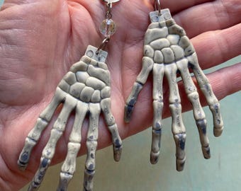 May include: A pair of off-white skeleton hand earrings featuring detailed bone structures with dark grey shading. Each earring is attached to a silver-toned metal leverback hook by a small jump ring and a single clear, faceted, iridescent bead.