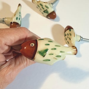 May include: A set of four ceramic cabinet knobs shaped like fish. Each knob features a cream-colored body with green painted spots and a distinct brown head. A silver-toned metal bolt extends from the back of each fish-shaped handle for mounting.