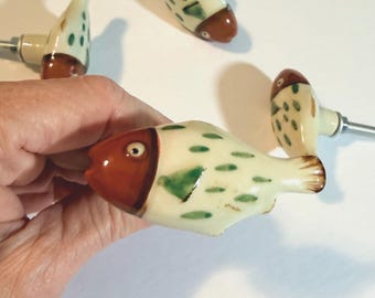 May include: A set of four ceramic cabinet knobs shaped like fish. Each knob features a cream-colored body with green painted spots and a distinct brown head. A silver-toned metal bolt extends from the back of each fish-shaped handle for mounting.