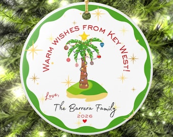 May include: A round white ceramic ornament with a green scalloped border. It features a palm tree decorated with lights and ornaments on a small island. Text reads: WARM WISHES FROM KEY WEST! Love, The Barrera Family 2026 with a red heart.