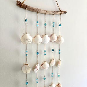 May include: A wall hanging featuring a piece of driftwood suspended by a brown cord. Five vertical strands of twine hang from the wood, each decorated with white scallop shells and small, translucent blue glass beads arranged in a pattern.