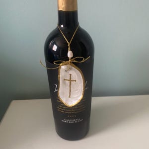 Puede incluir: Una botella de vino tinto con etiqueta negra y un adorno colgante. El adorno consiste en una concha de ostra blanca con una cruz dorada, una peque&ntilde;a perla blanca y un cord&oacute;n met&aacute;lico dorado atado en forma de lazo alrededor del cuello.