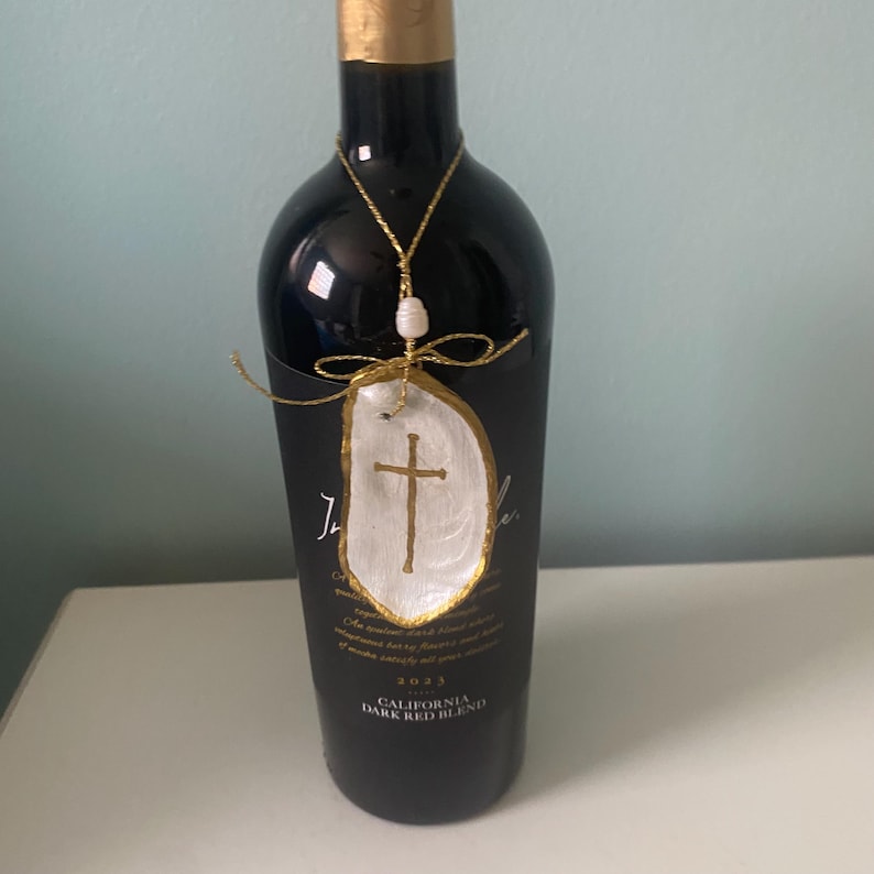 Puede incluir: Una botella de vino tinto con etiqueta negra y un adorno colgante. El adorno consiste en una concha de ostra blanca con una cruz dorada, una peque&ntilde;a perla blanca y un cord&oacute;n met&aacute;lico dorado atado en forma de lazo alrededor del cuello.