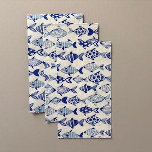 May include: Three rectangular paper napkins with a repeating pattern of blue fish on a white background. The fish designs vary, including stripes, dots, and solid blue. The napkins are arranged in a slightly overlapping manner.