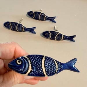 May include: Four fish-shaped cabinet knobs featuring a glossy dark blue and white finish. Each knob has a detailed scale pattern on the body, a defined eye, and a metal screw post extending from the back for attachment to furniture or cabinetry.