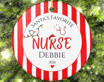 May include: A round ceramic Christmas ornament with red and white vertical stripes on the border. The center features the text "SANTA'S FAVORITE NURSE DEBBIE 2026", a red stethoscope, a string of colorful Christmas lights, and a Santa hat.