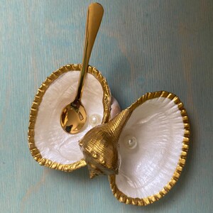 May include: Two white seashell dishes with gold-painted edges, each containing a small white pearl. A gold-toned metal spoon rests in one shell, and a gold-painted spiral seashell is positioned between the two larger shells.