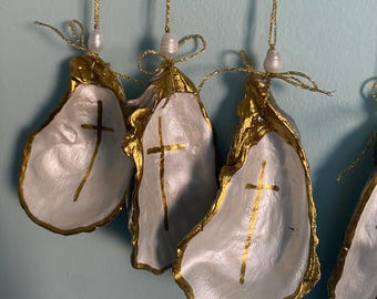 May include: A set of hanging oyster shell ornaments, each featuring a white interior painted with a gold cross. The edges are gilded in gold, and each shell is topped with a small white pearl bead and a thin gold glitter string for hanging.