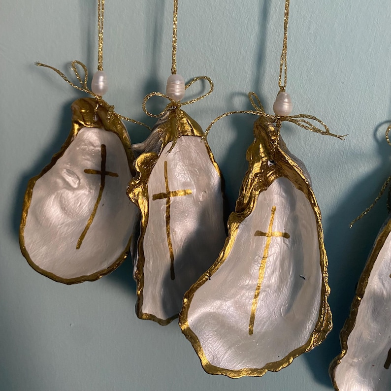 Puede incluir: Un conjunto de adornos colgantes de conchas de ostra, cada una con un interior blanco pintado con una cruz dorada. Los bordes est&aacute;n dorados y cada concha est&aacute; rematada con una peque&ntilde;a perla blanca y un cord&oacute;n dorado brillante para colgar.