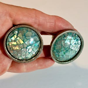 May include: Two circular cabinet knobs featuring a silver-toned metal frame and a decorative inlay of iridescent, mosaic-style shell fragments in shades of teal, blue, and white. The surface of the inlay is smooth, glossy, and reflective.