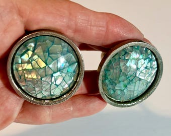 May include: Two circular cabinet knobs featuring a silver-toned metal frame and a decorative inlay of iridescent, mosaic-style shell fragments in shades of teal, blue, and white. The surface of the inlay is smooth, glossy, and reflective.