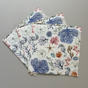 May include: A stack of decorative paper napkins with a sea life design. The napkins feature illustrations of seahorses, starfish, coral, and seashells in shades of blue, red, and beige on a white background.