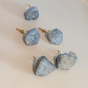 May include: Five cabinet knobs featuring irregular, rough-cut light blue druzy stone faces with sparkling crystalline textures. Each knob is attached to a gold-toned metal post and base, providing a functional handle for furniture or cabinetry.