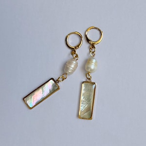 May include: A pair of gold-colored drop earrings. Each earring features a rectangular, iridescent mother-of-pearl charm with a gold border, connected to a freshwater pearl and a gold hoop. The earrings are displayed on a white background.