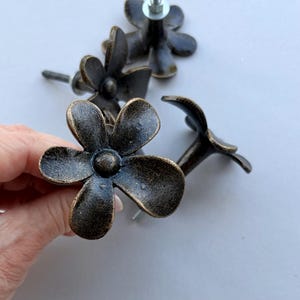 May include: A set of three dark bronze-toned metal drawer knobs, each shaped like a five-petaled flower with a central circular detail. The knobs feature a textured, weathered finish and include a threaded metal bolt extending from the back.